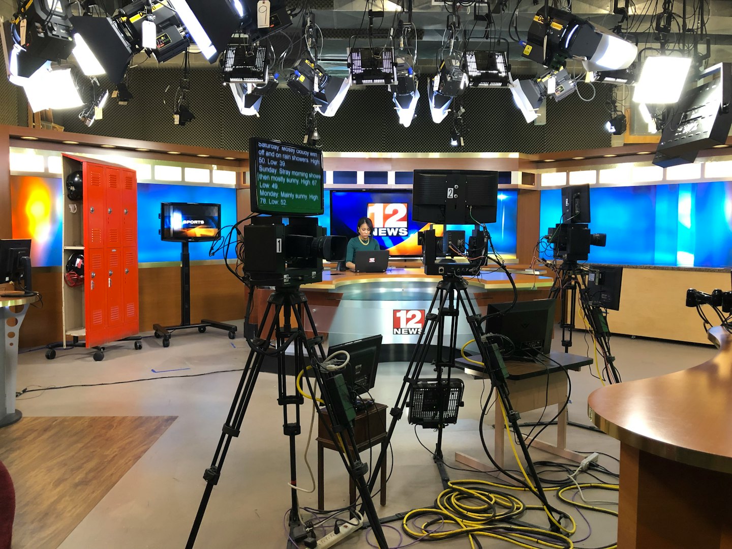 Person sits at news desk with cameras and weather screens.